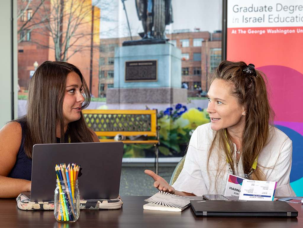two students face each other behind desk, laptop open