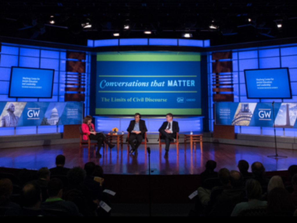Three panelists on stage with large screens in backround that say "Conversations that Matter" and the GW logo